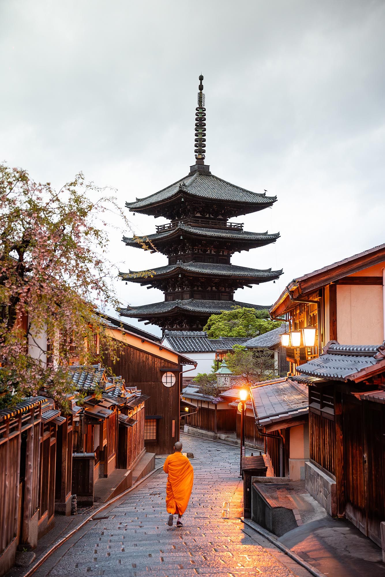 Historic Kyoto street with traditional wooden buildings and pagoda illuminated at dusk with orange-robed figure walking.