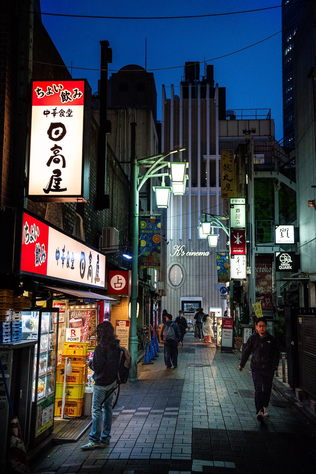 Illuminated Japanese street at night with neon signs and silhouettes of people walking along a narrow alley in Tokyo.