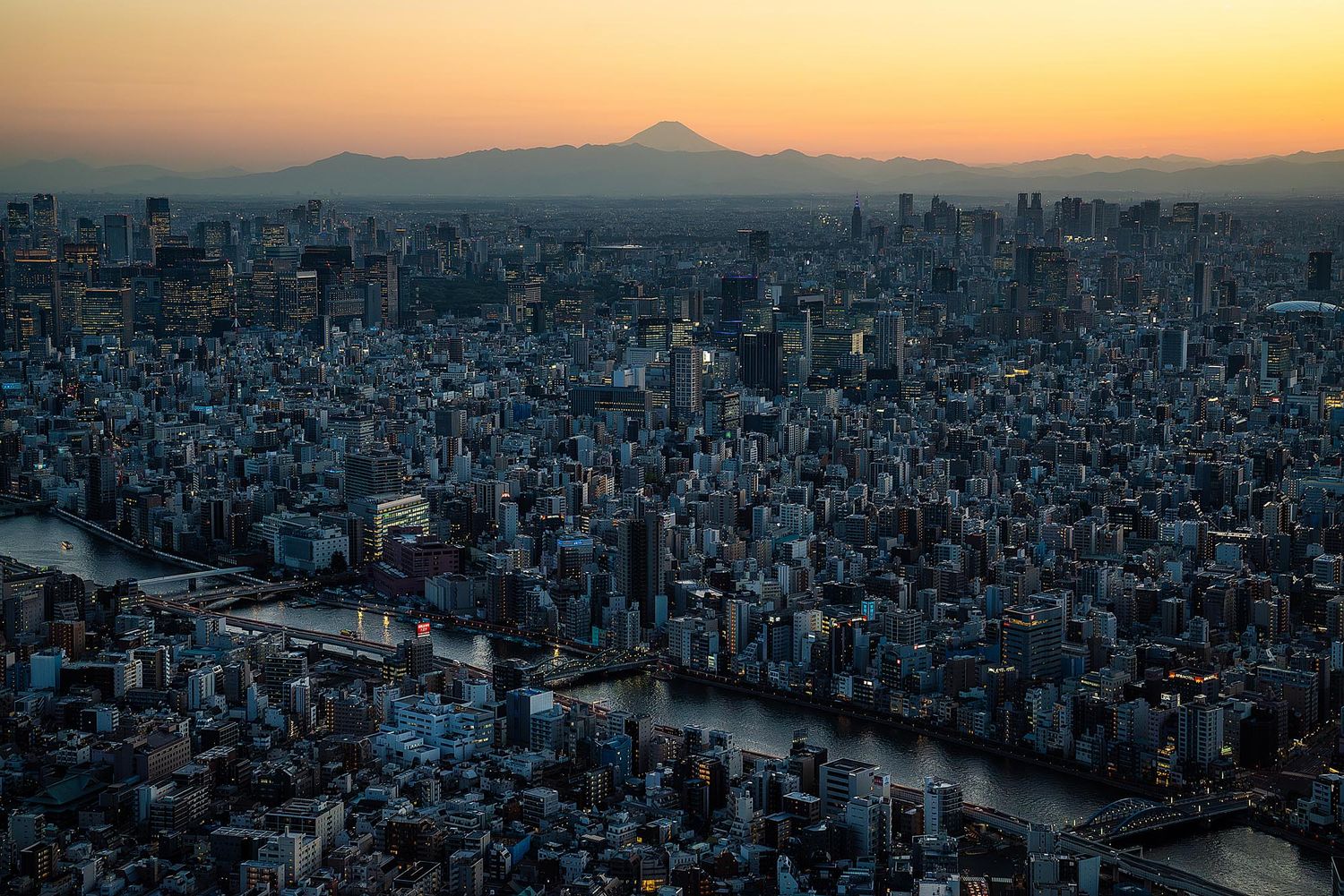 Panoramic aerial view of Tokyo cityscape at sunset with Mount Fuji silhouetted in the background.
