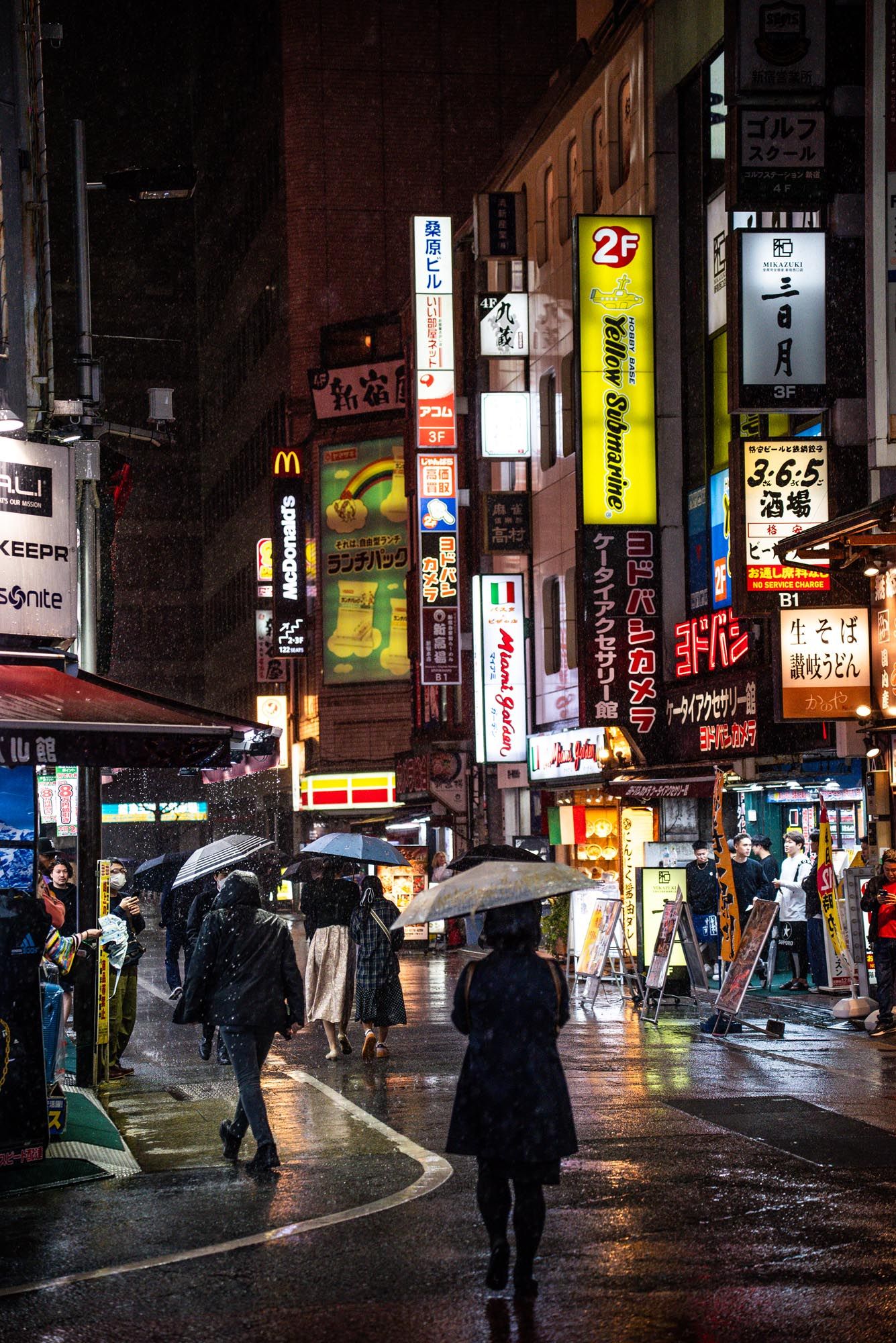Pedestrians with umbrellas walk through a rainy night street in Tokyo with bright neon signs and advertisements.