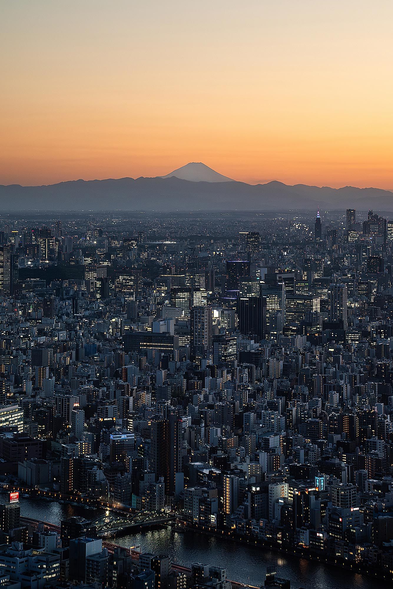 Panoramic view of Tokyo cityscape at sunset with Mount Fuji silhouetted against an orange sky.