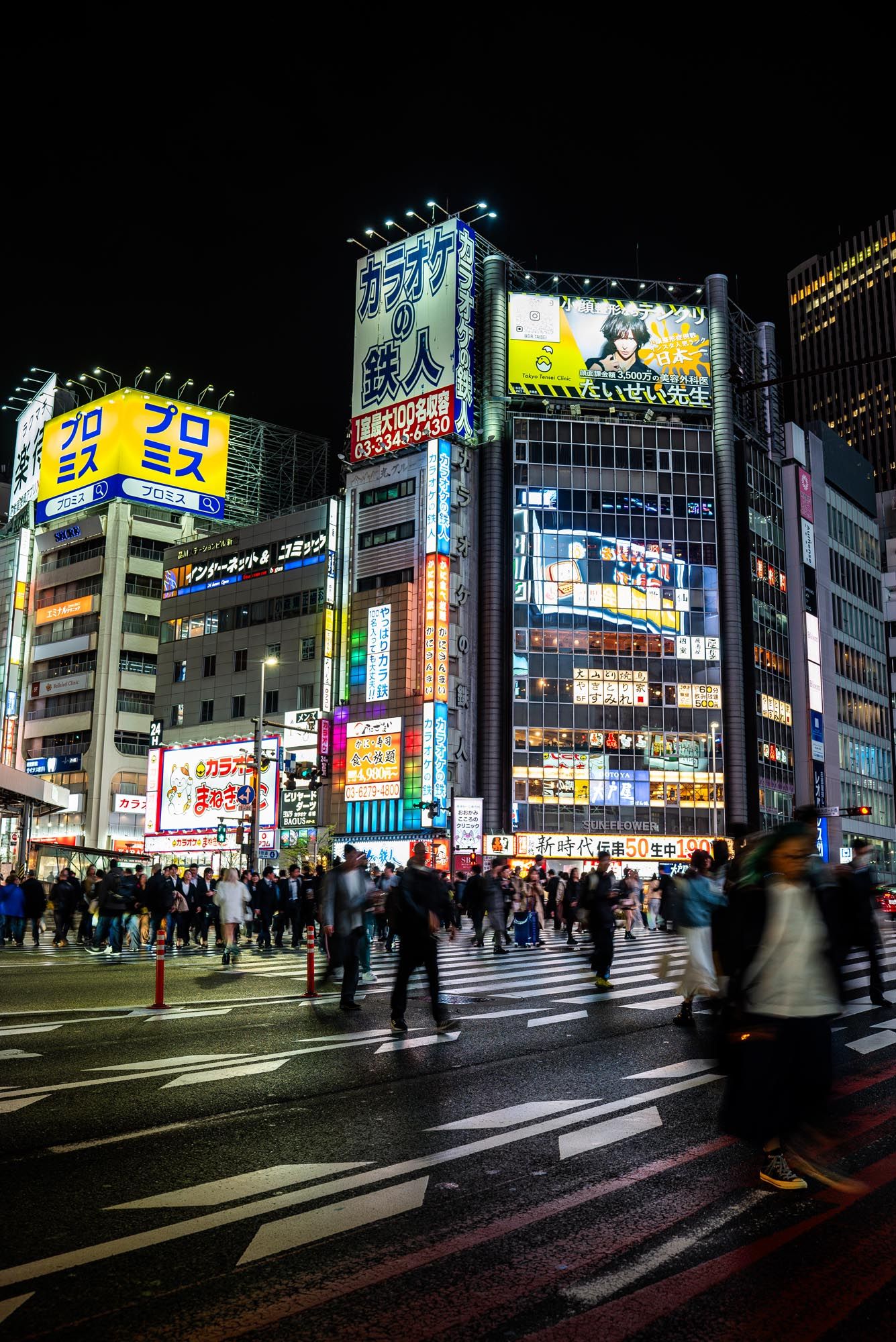 Time-lapse panorama of bustling Shinjuku district at night with bright neon signs and blurred pedestrians crossing street.