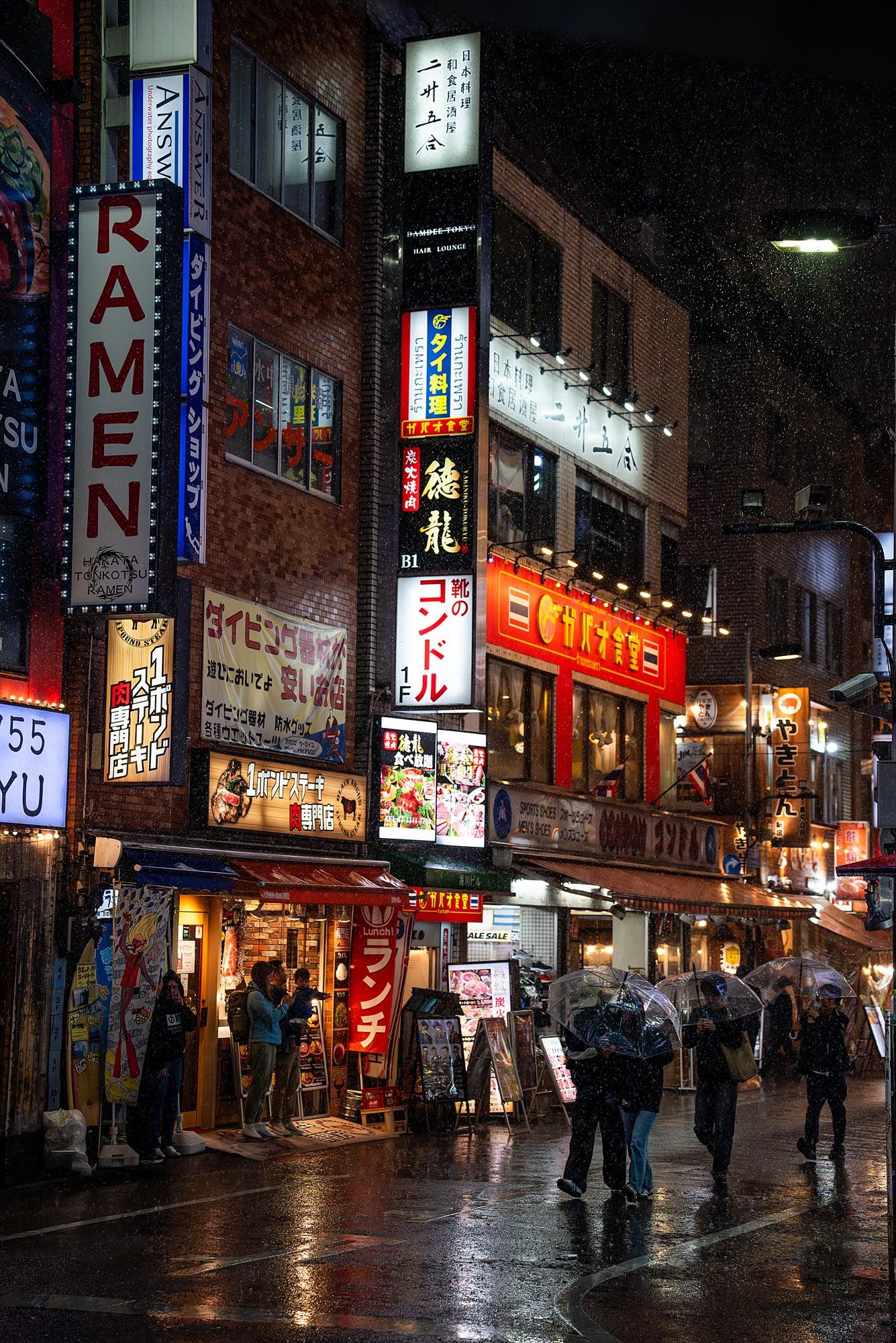Neon signs illuminate a rainy night street scene in Japan's entertainment district with pedestrians walking under umbrellas.