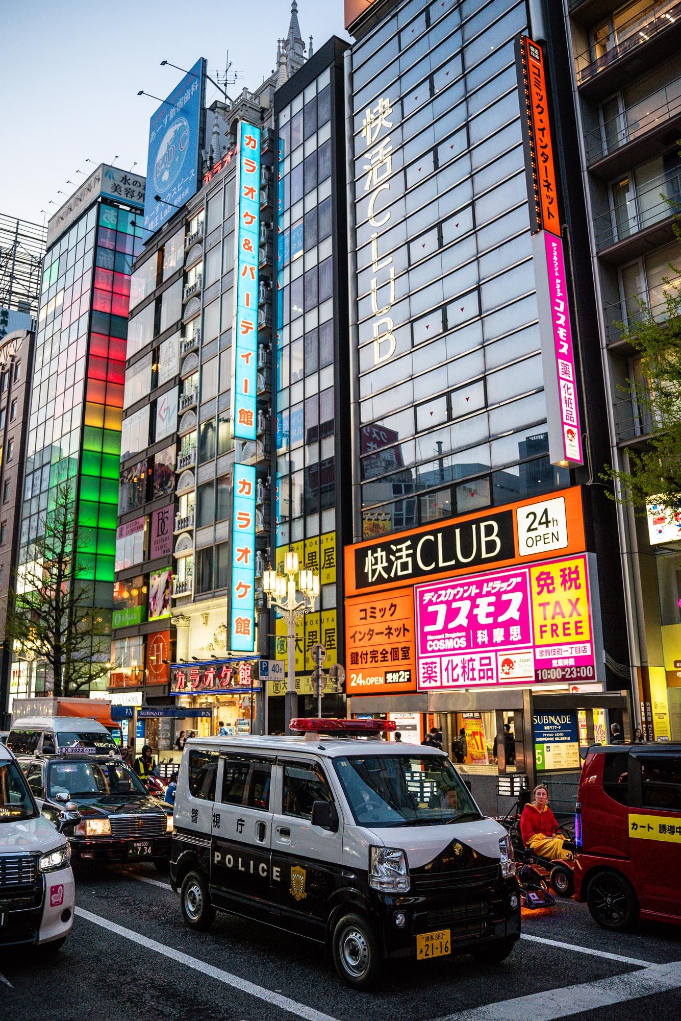 Colorful neon signs and modern buildings line a street in Tokyo's bustling Akihabara district with police cars parked nearby.