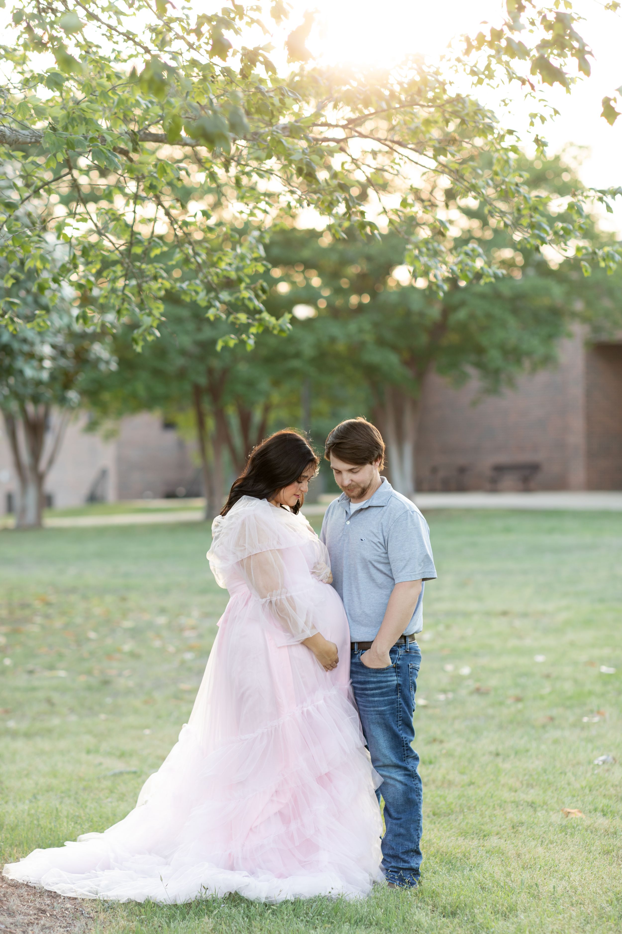 Romantic outdoor maternity photo with couple standing together at sunset in grassy field.