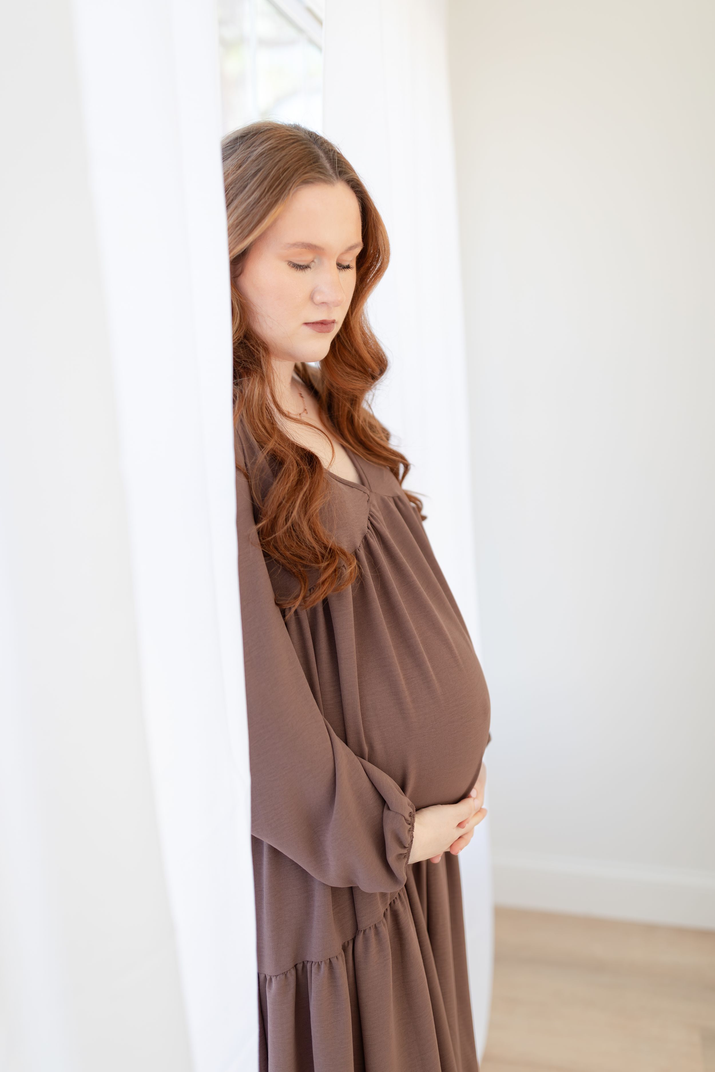 Expectant mother in brown flowing dress poses gracefully by window in soft natural light.
