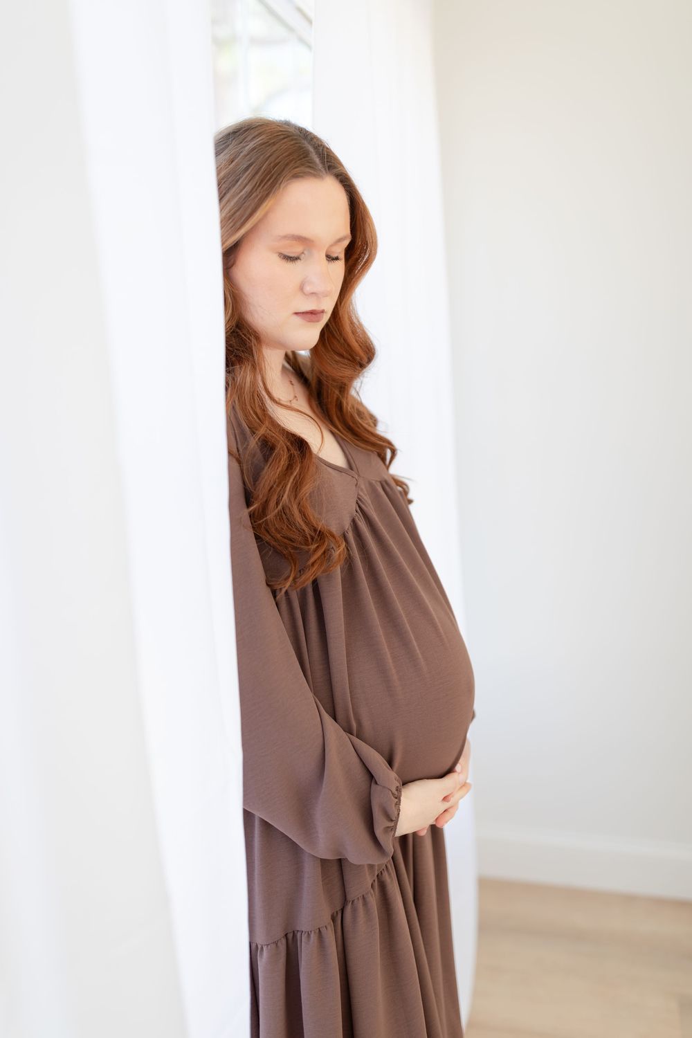 Expectant mother in brown flowing dress poses gracefully by window in soft natural light.