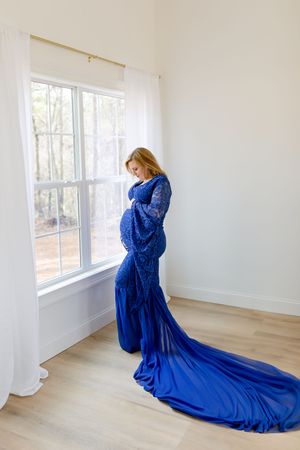 Elegant maternity photo featuring flowing blue gown with long train by sunlit window.