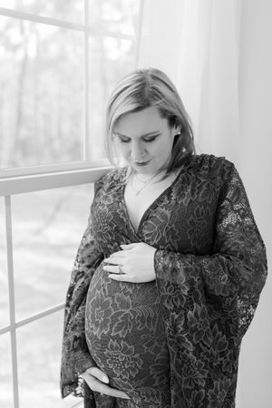 Maternity portrait in black and white showing woman in lace dress by bright window.