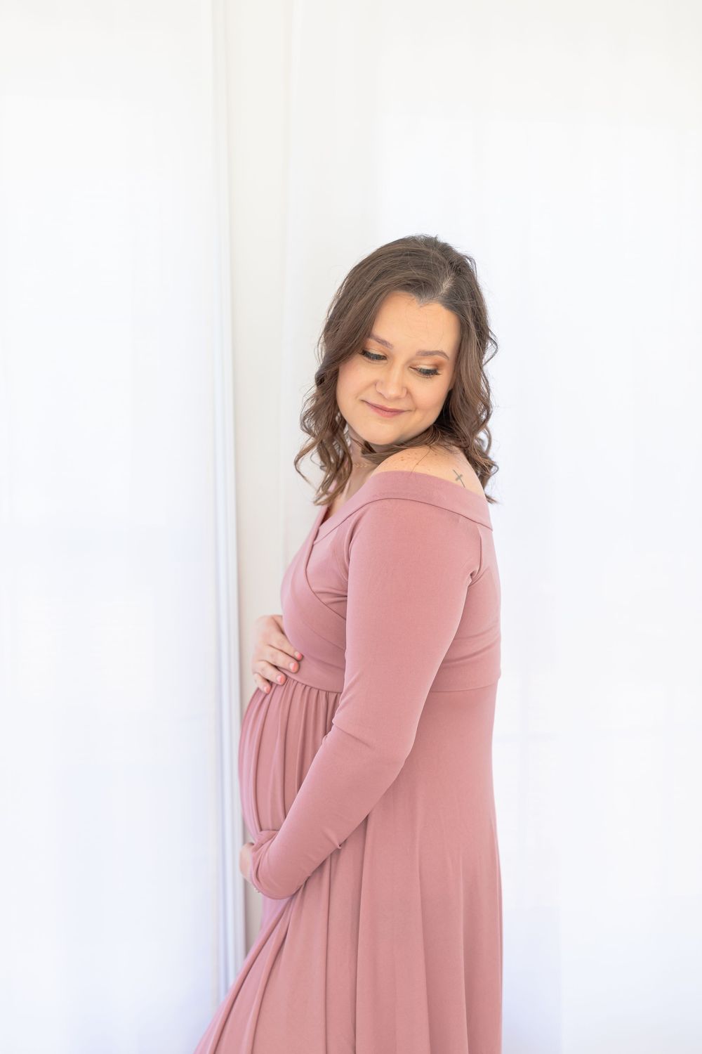 Maternity portrait in dusty rose dress against white wall with natural lighting.