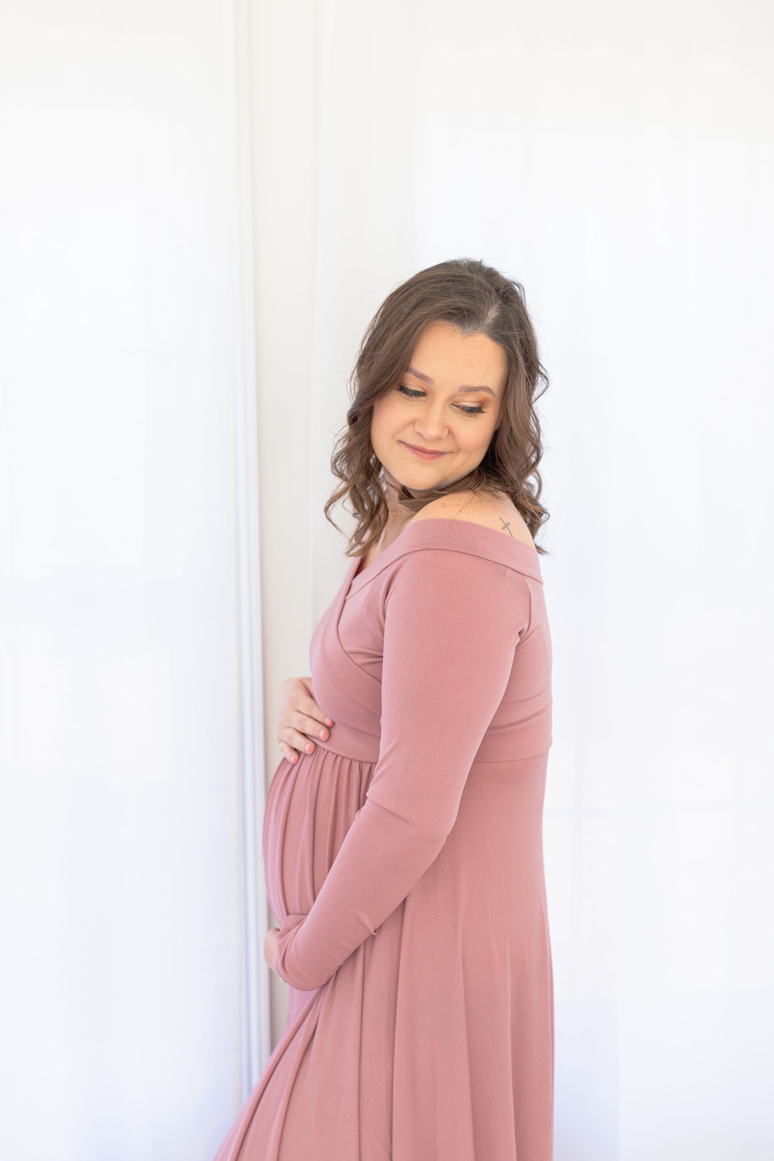 Maternity portrait in dusty rose dress against white wall with natural lighting.