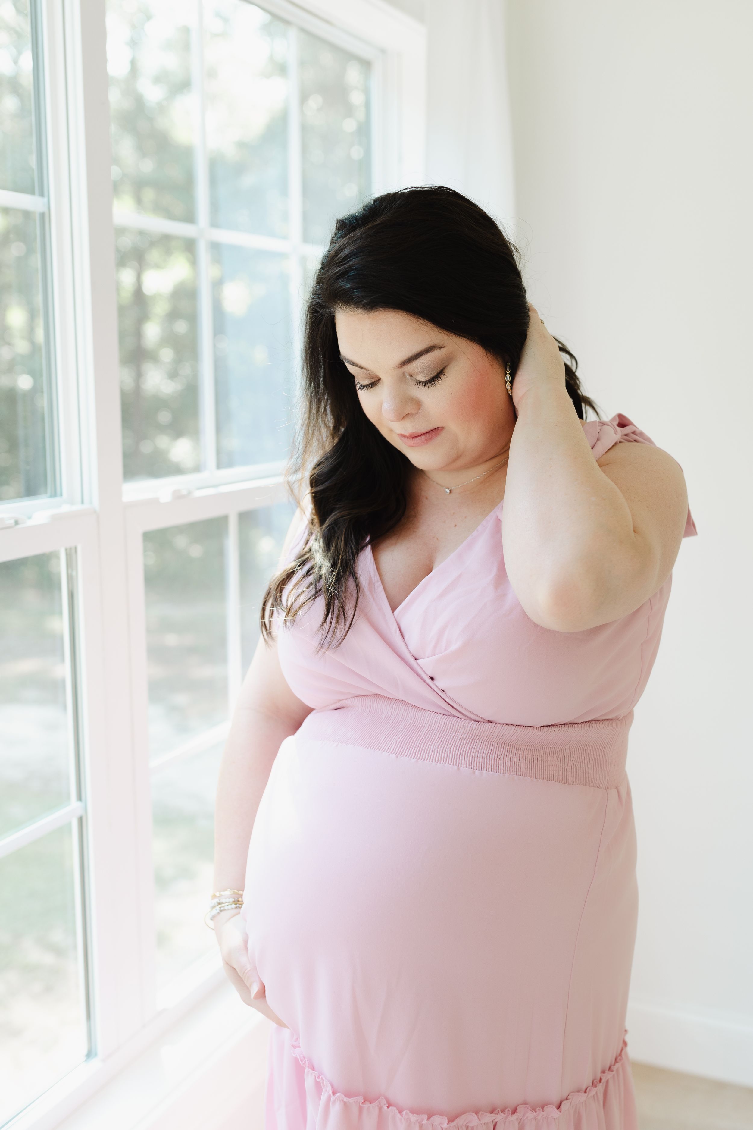 Intimate maternity portrait in pink dress by window with soft natural light.