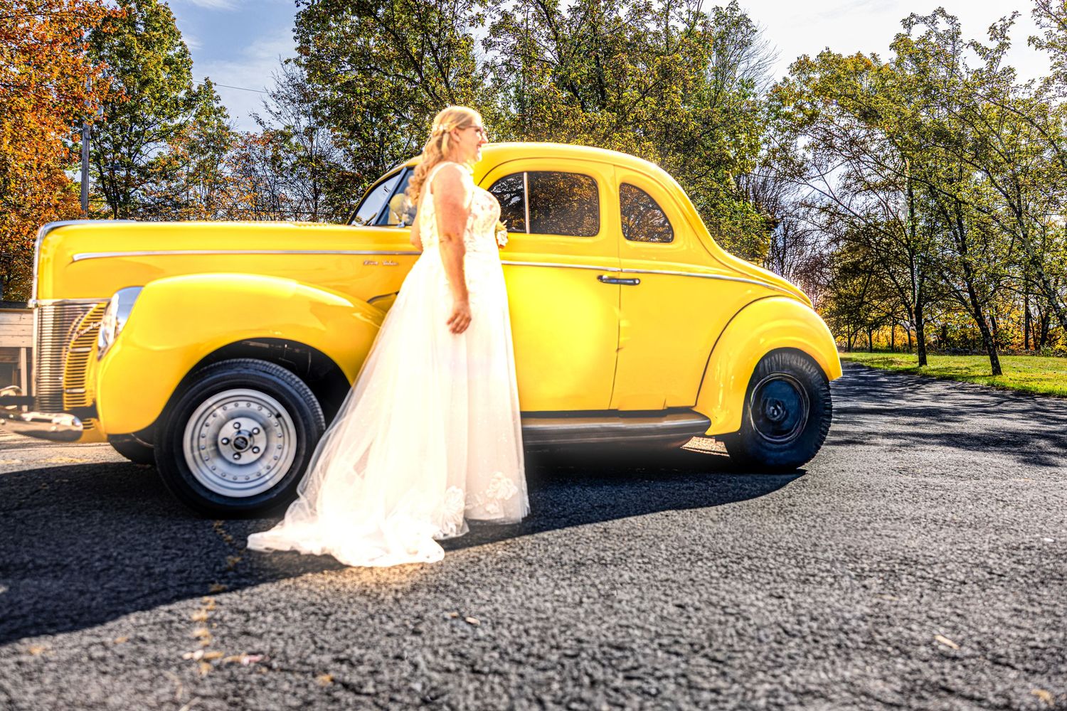 A bright yellow vintage car parked outdoors with someone in formal attire standing beside it on a sunny autumn day.