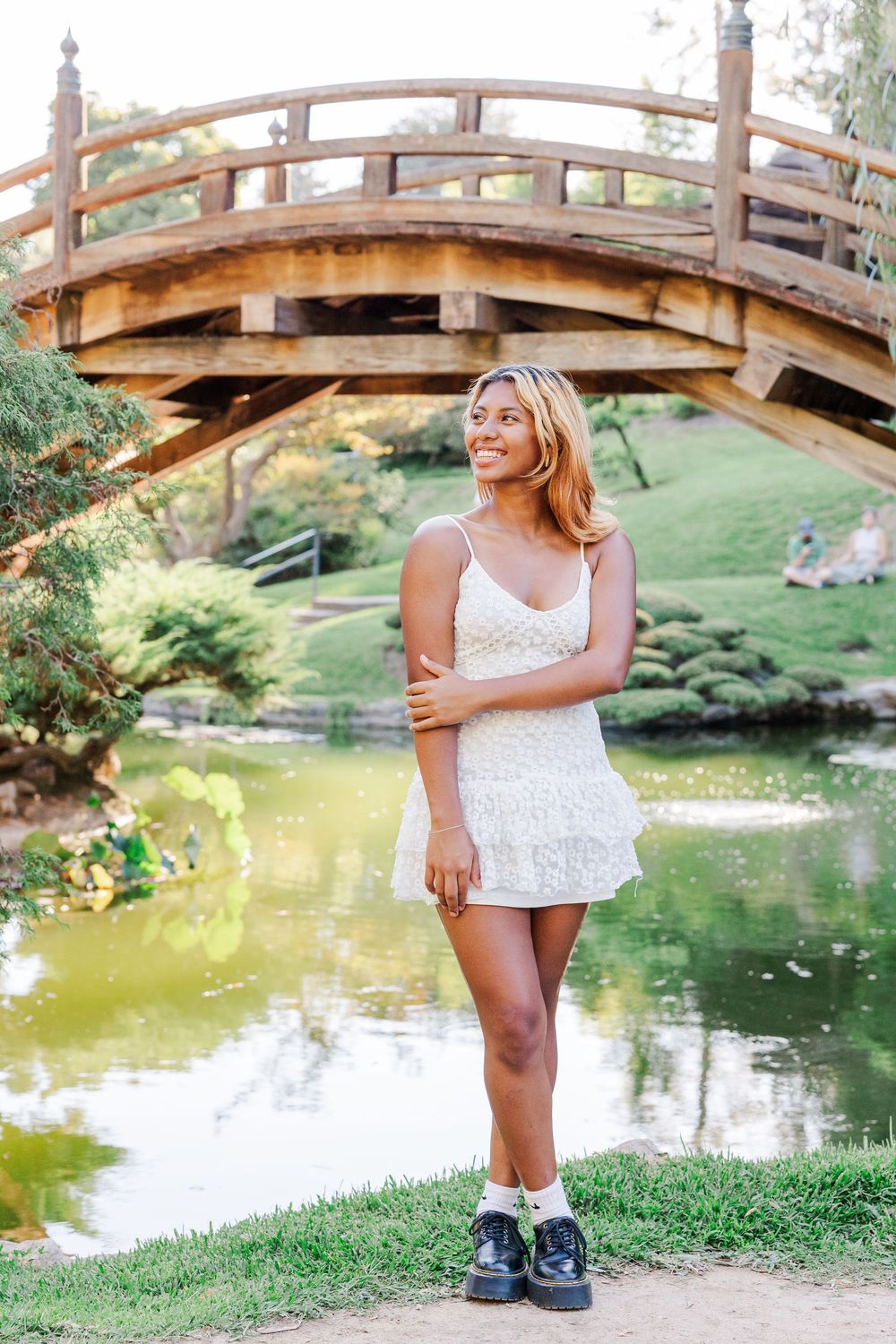 A wooden footbridge arches over a serene pond in a peaceful garden setting with lush greenery.