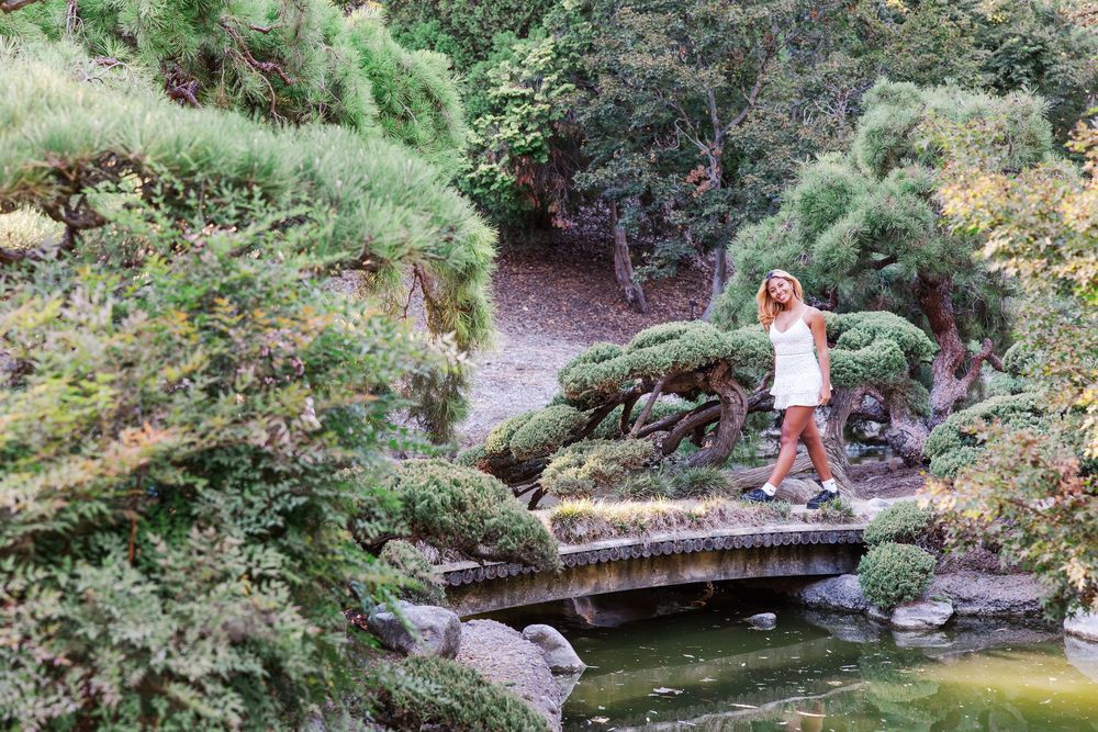 A person crosses a wooden footbridge over a tranquil pond in a lush Japanese garden setting.