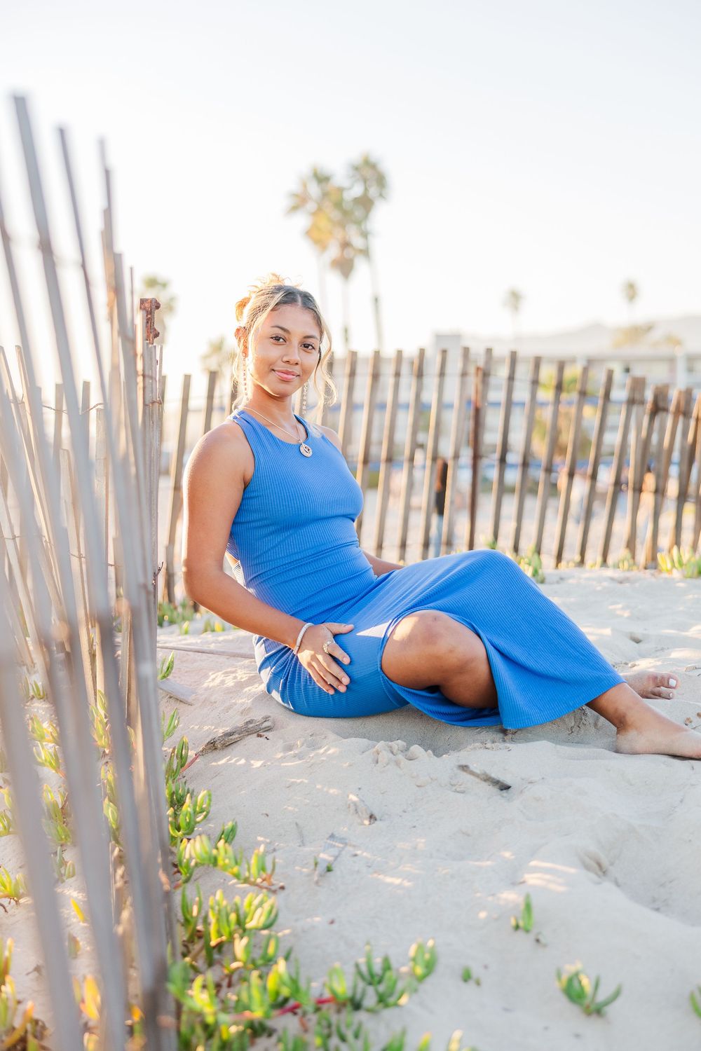 Woman in blue dress relaxes on sandy beach with wooden fence and palm trees at sunset.