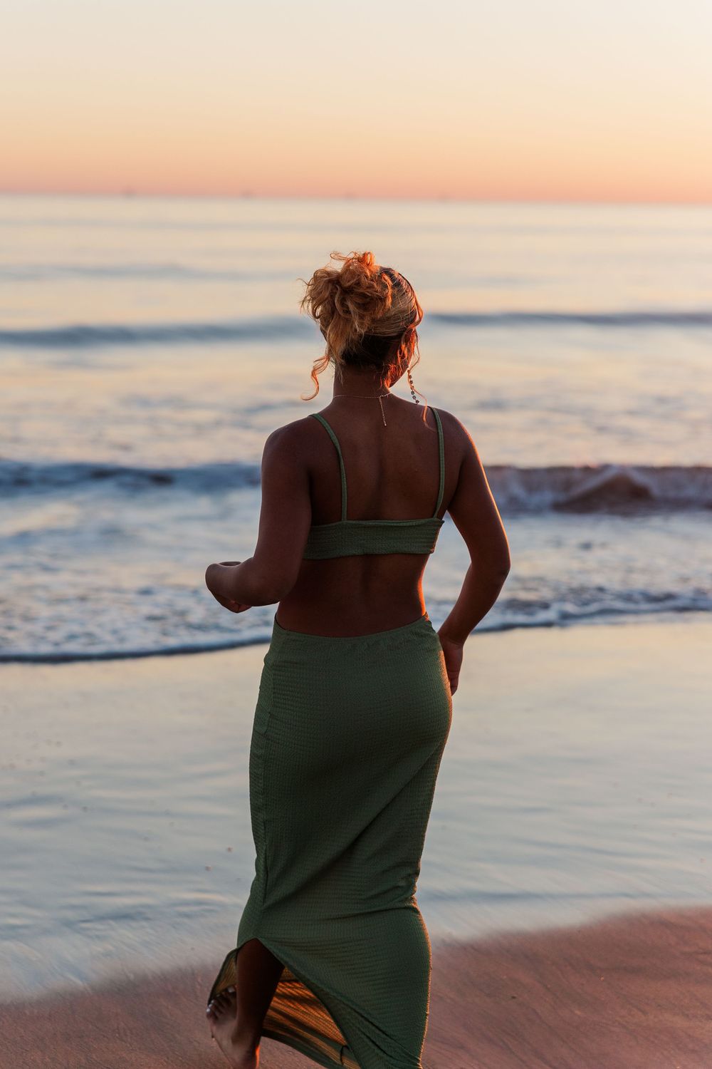 Figure in green dress walks along ocean shore at sunset with waves crashing behind.