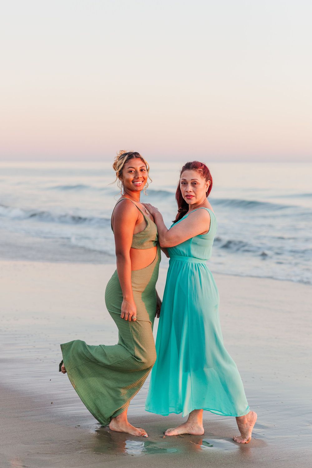 Two people in green and turquoise dresses pose together on beach at sunset.