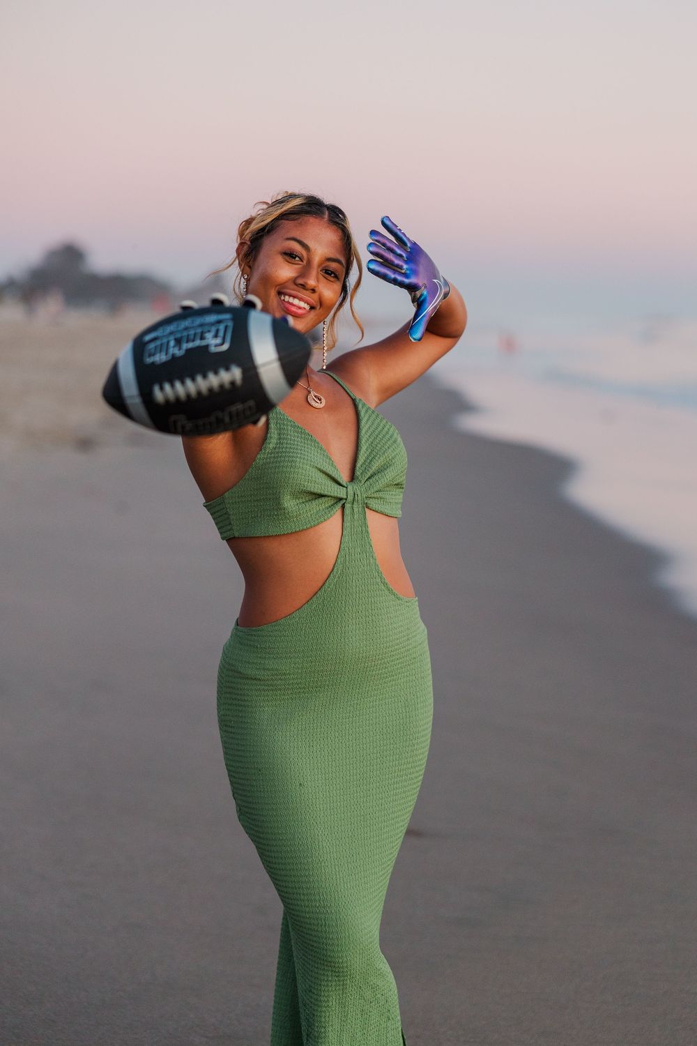 Woman in green bodycon dress poses playfully on beach at sunset with water bottle.