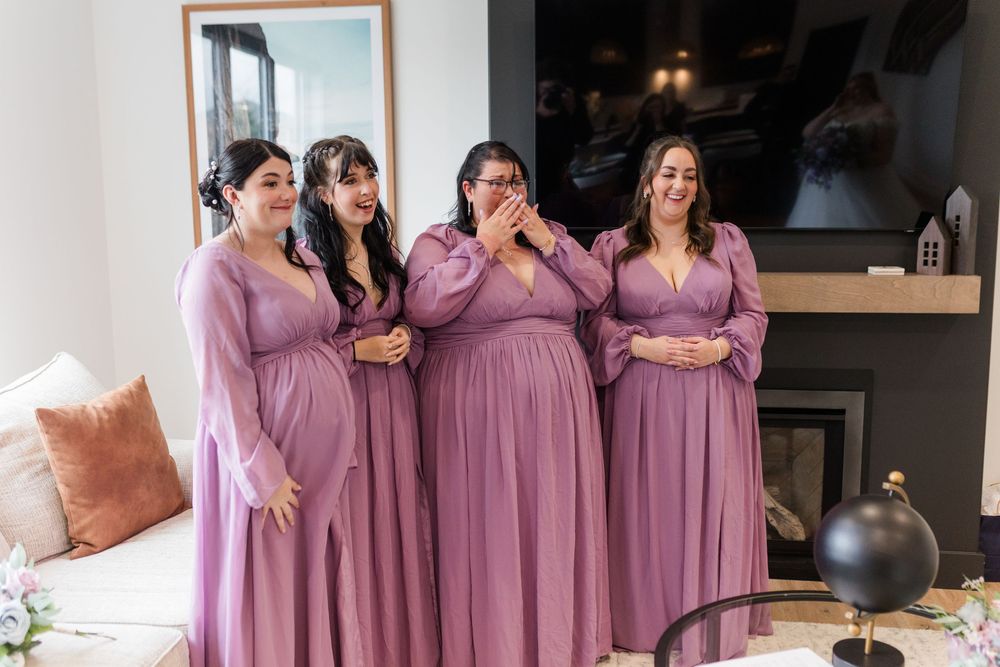 Bridesmaids in dusty rose dresses gather together for wedding party photos in a bright indoor setting.