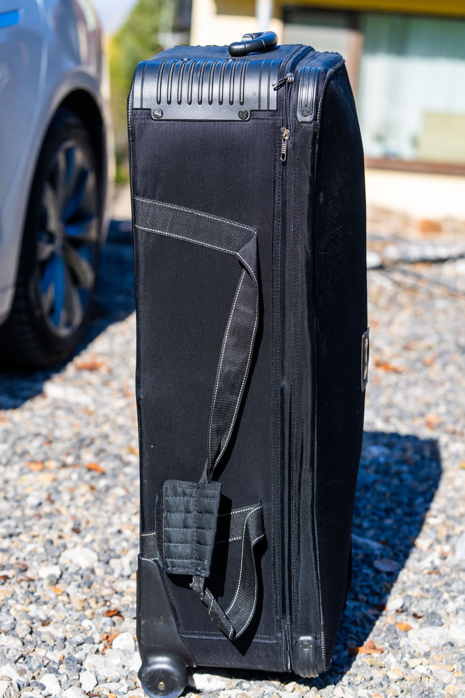 Black hard shell golf travel case with wheels and handle standing on gravel next to a parked car.
