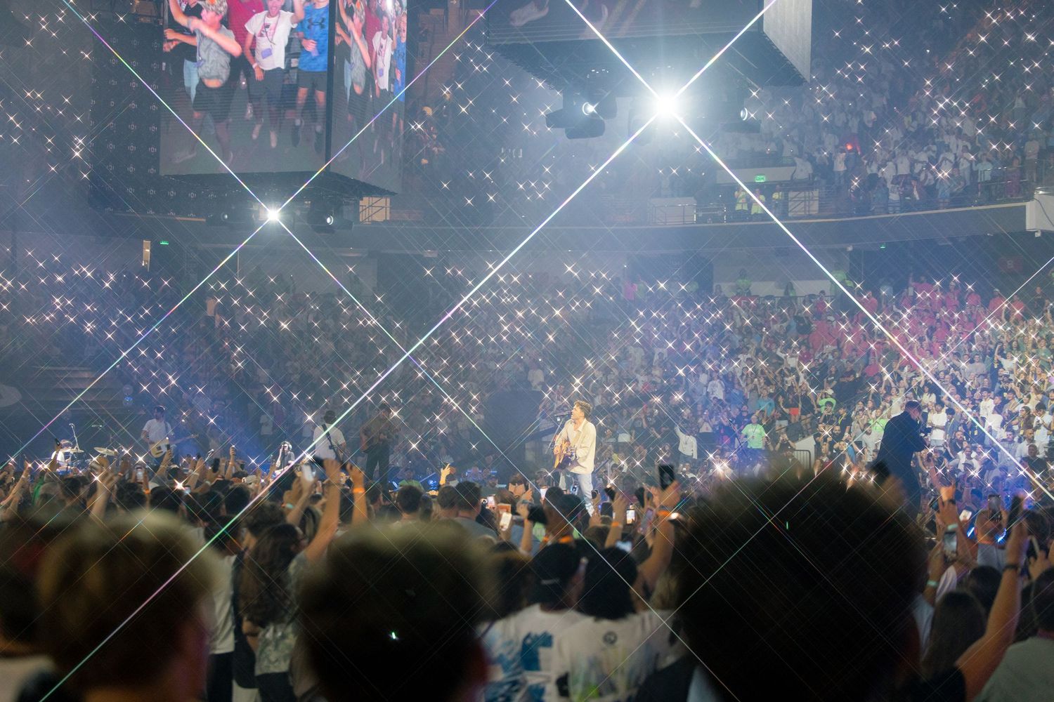 Concert crowd silhouetted against blue stage lights with text displayed on large screens.