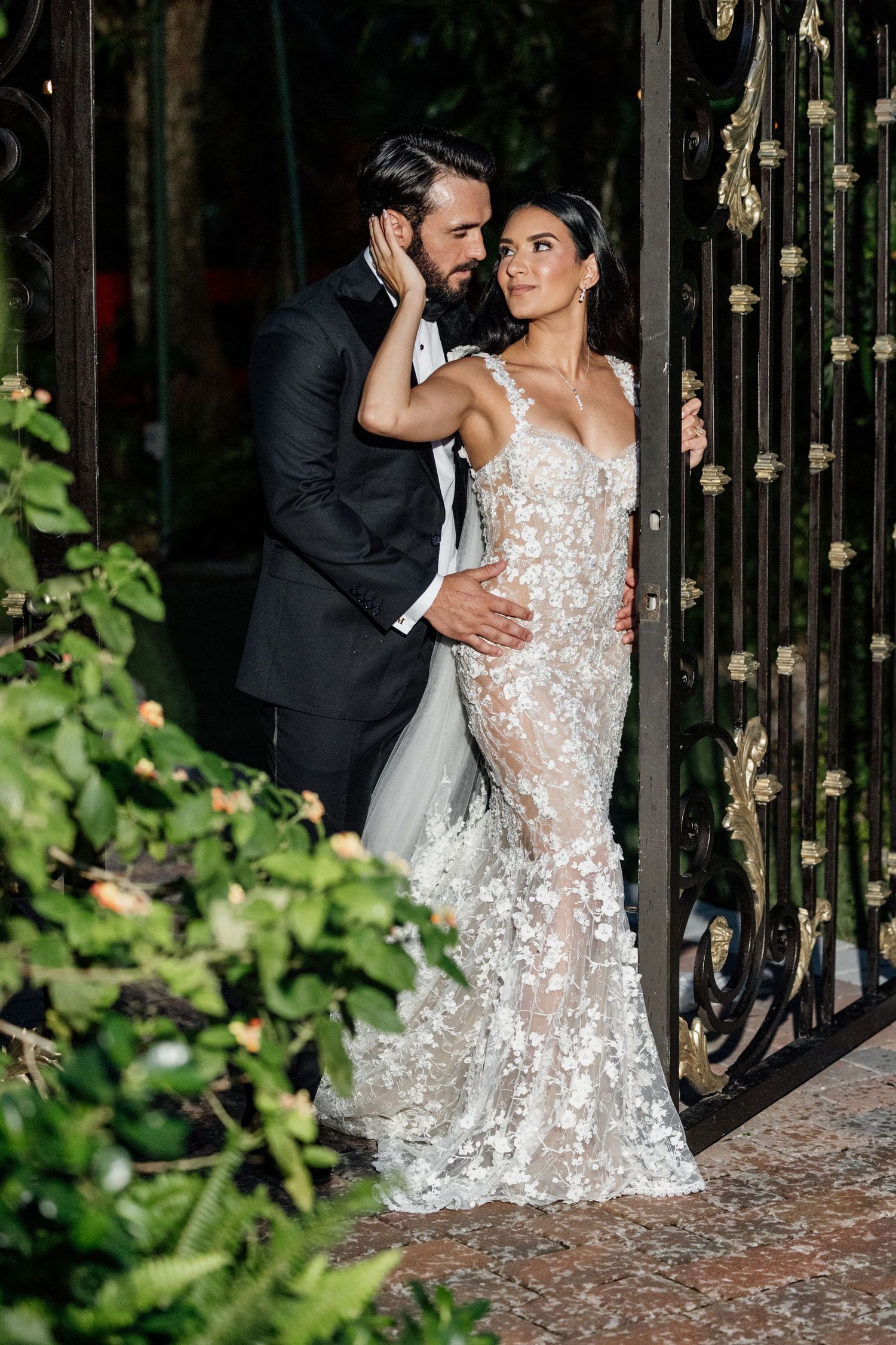 A romantic couple pose by an ornate garden gate in elegant wedding attire.