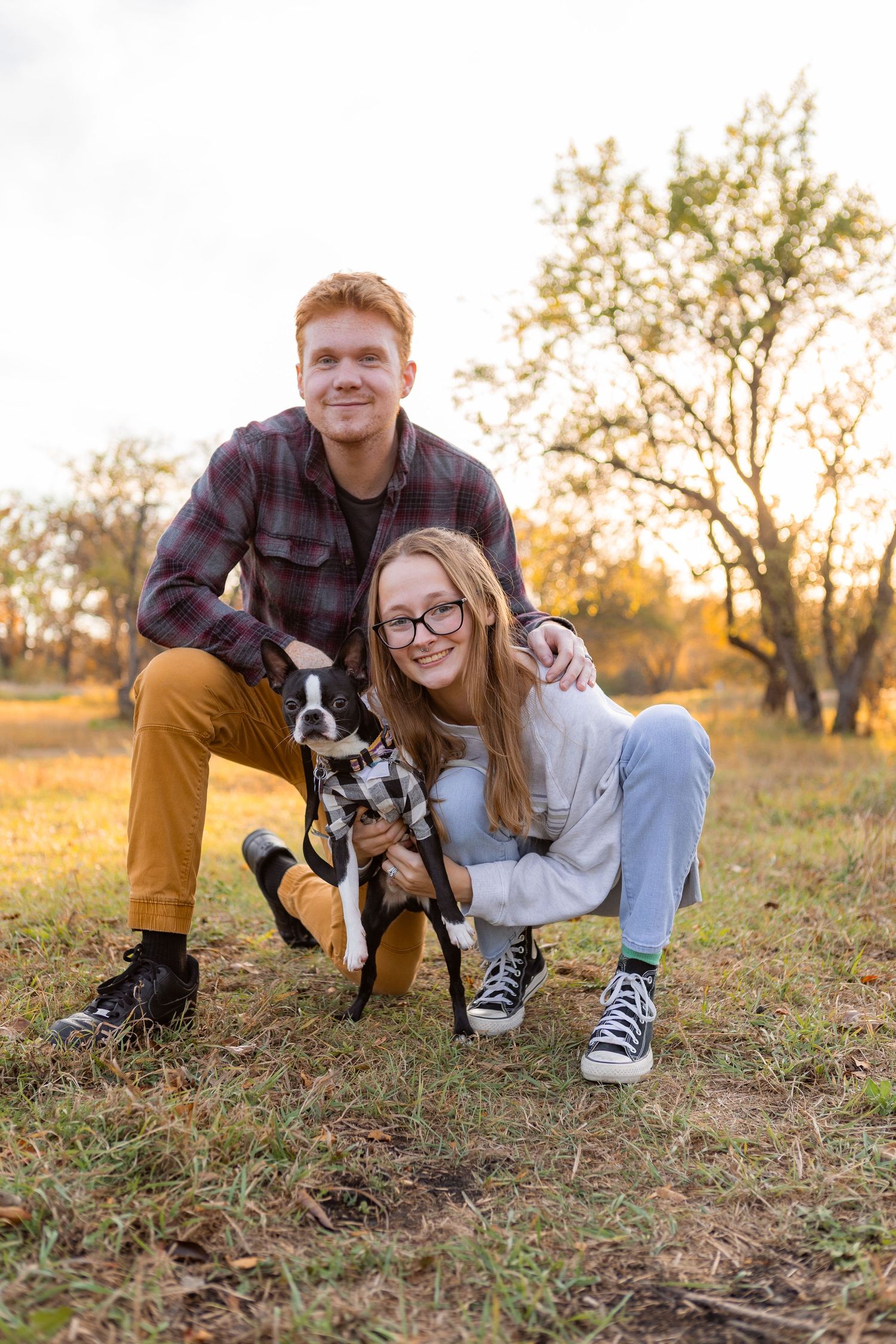 A couple sits in a grassy field at sunset with their black and white Boston Terrier.