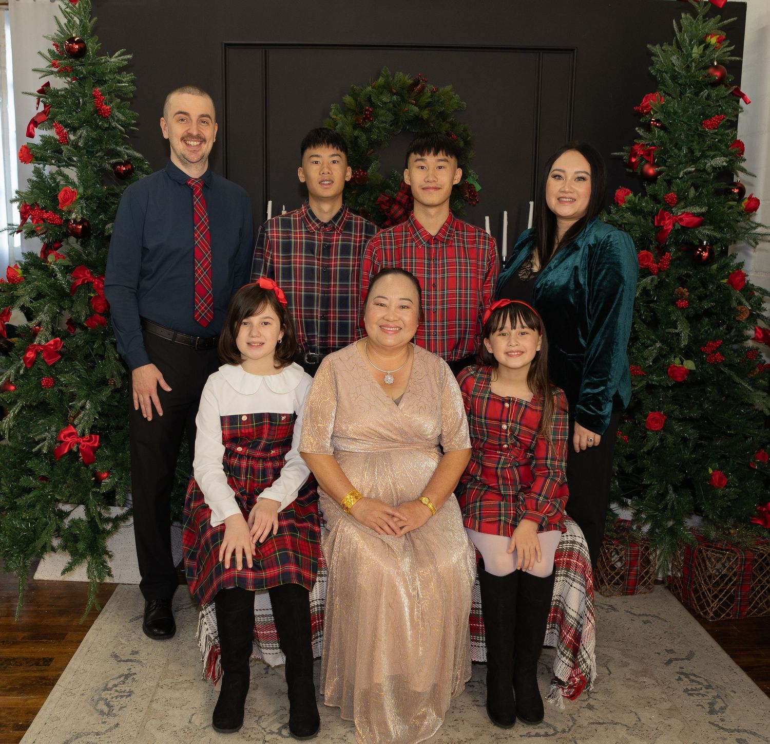 Family holiday portrait with coordinated red plaid outfits posed in front of decorated Christmas trees with red ornaments.