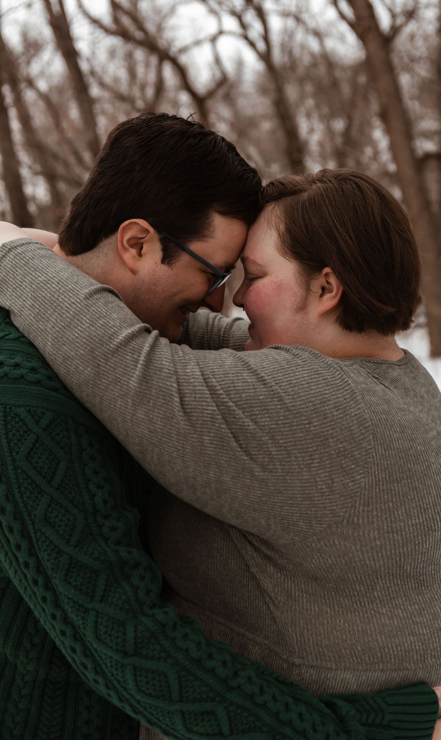 A tender romantic moment between a couple wearing cozy sweaters embracing in a winter forest setting.