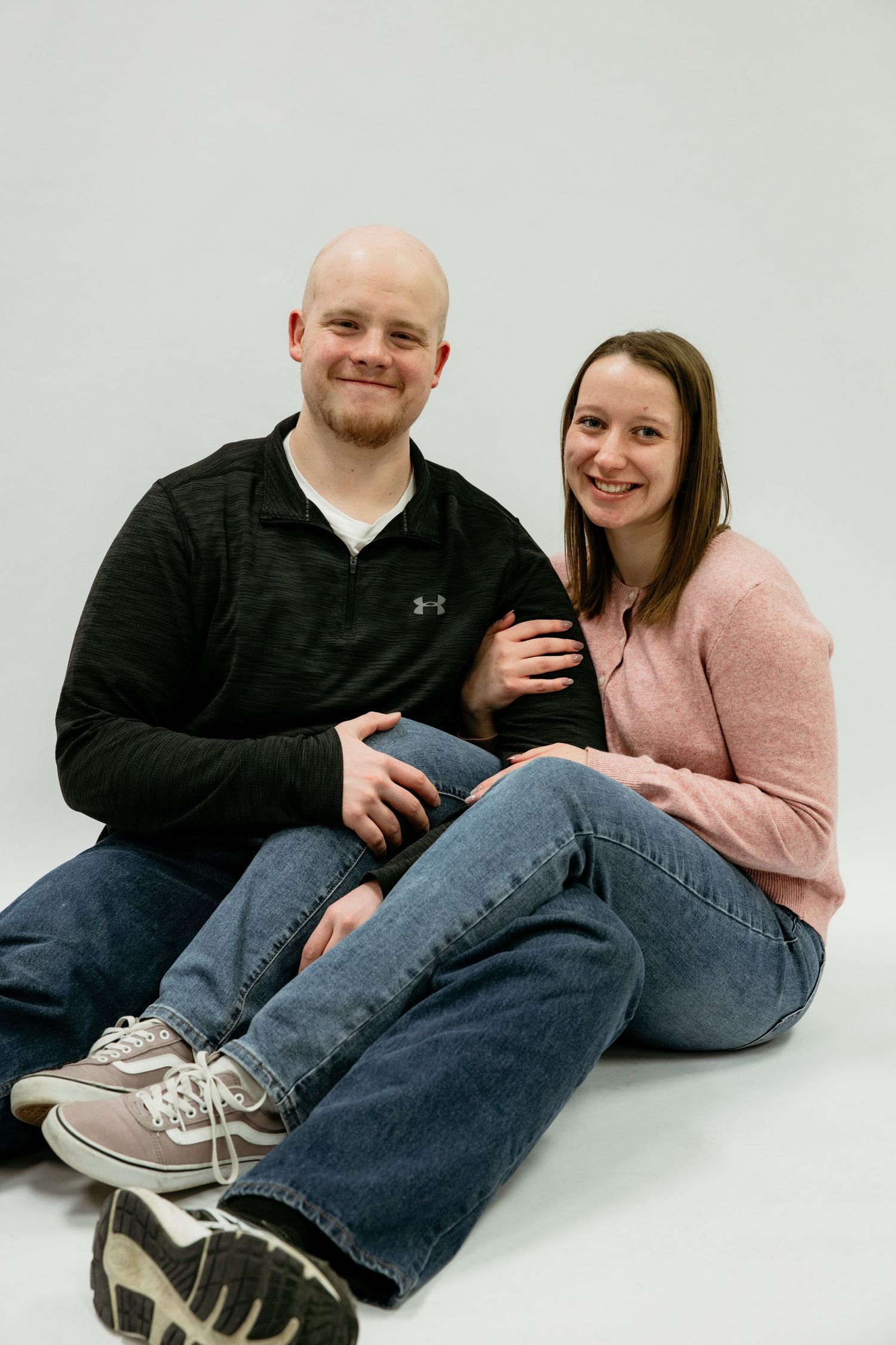 A couple in casual attire sits together on a white background for a portrait session.