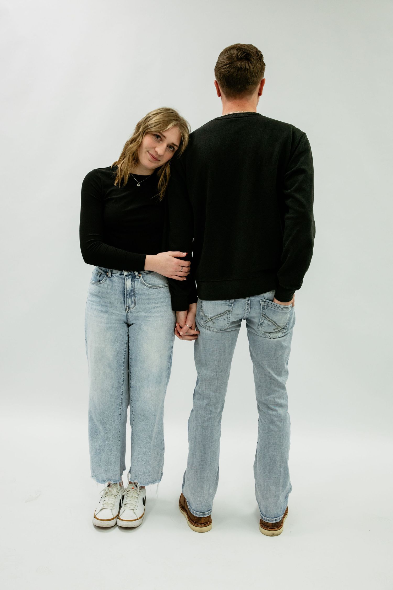 Two people wearing matching light wash denim jeans and contrasting tops stand together in a studio setting.