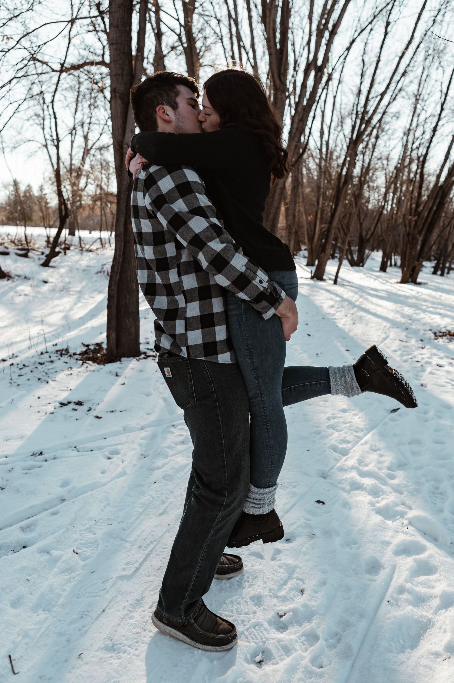 A couple shares a tender moment on a snowy winter path surrounded by bare trees in the forest.