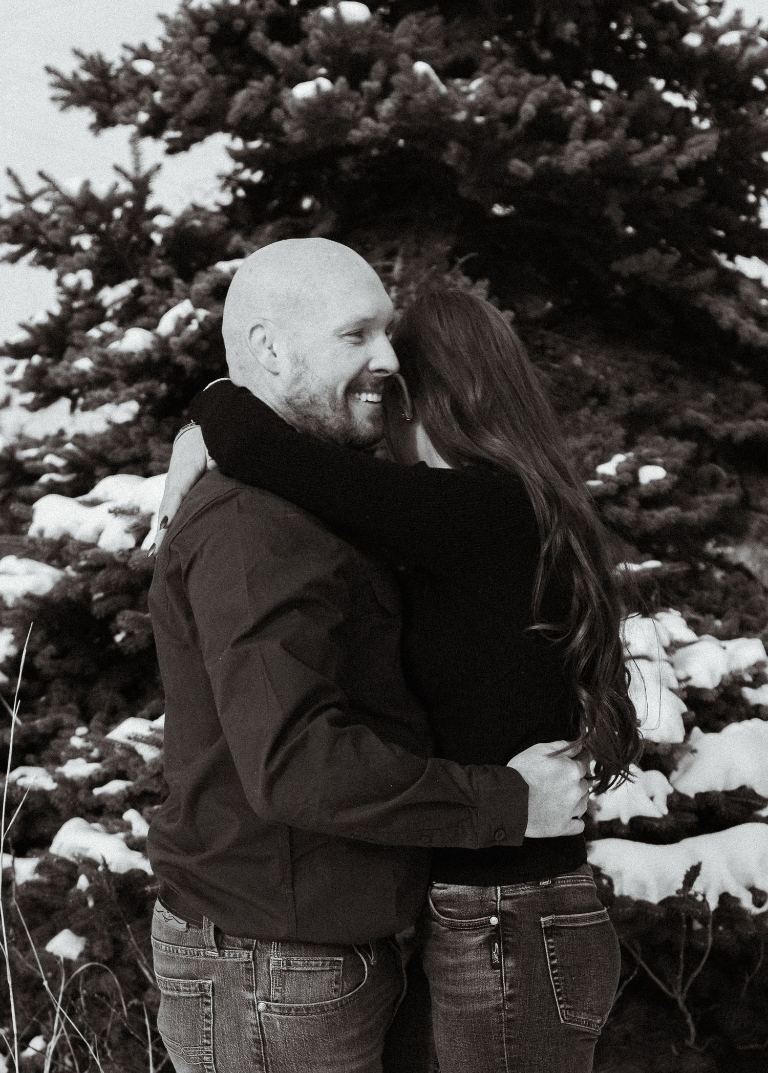 Black and white photo series showing a couple embracing in front of snow-covered evergreen trees in winter.
