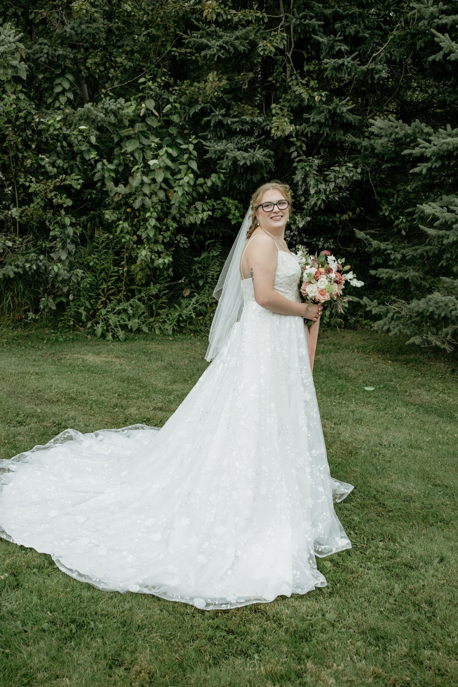 Bride in white lace gown with veil and floral bouquet posing outdoors on green lawn with trees.