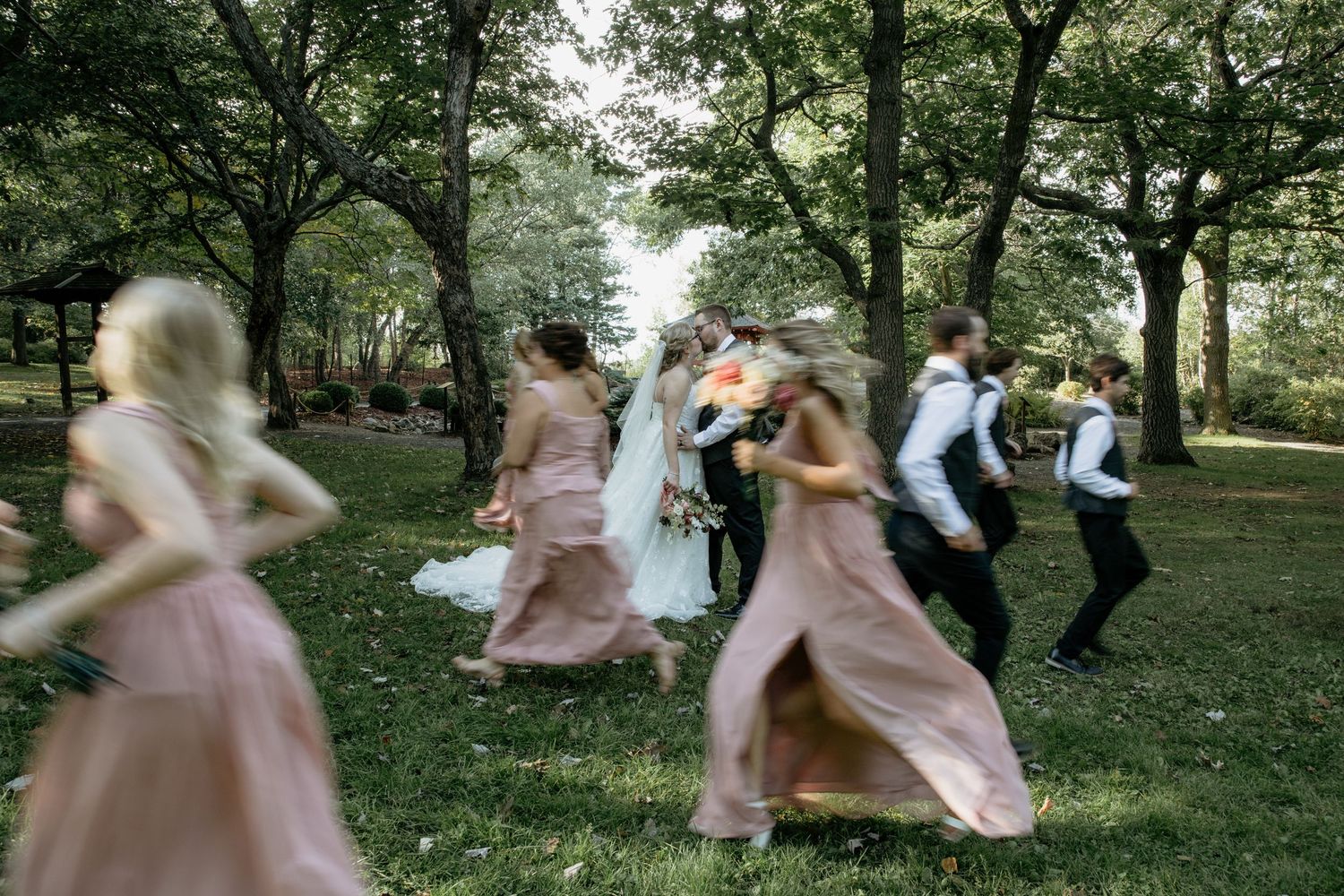 Wedding party in motion outdoors, bridesmaids in blush pink gowns and groomsmen celebrating among lush green trees.