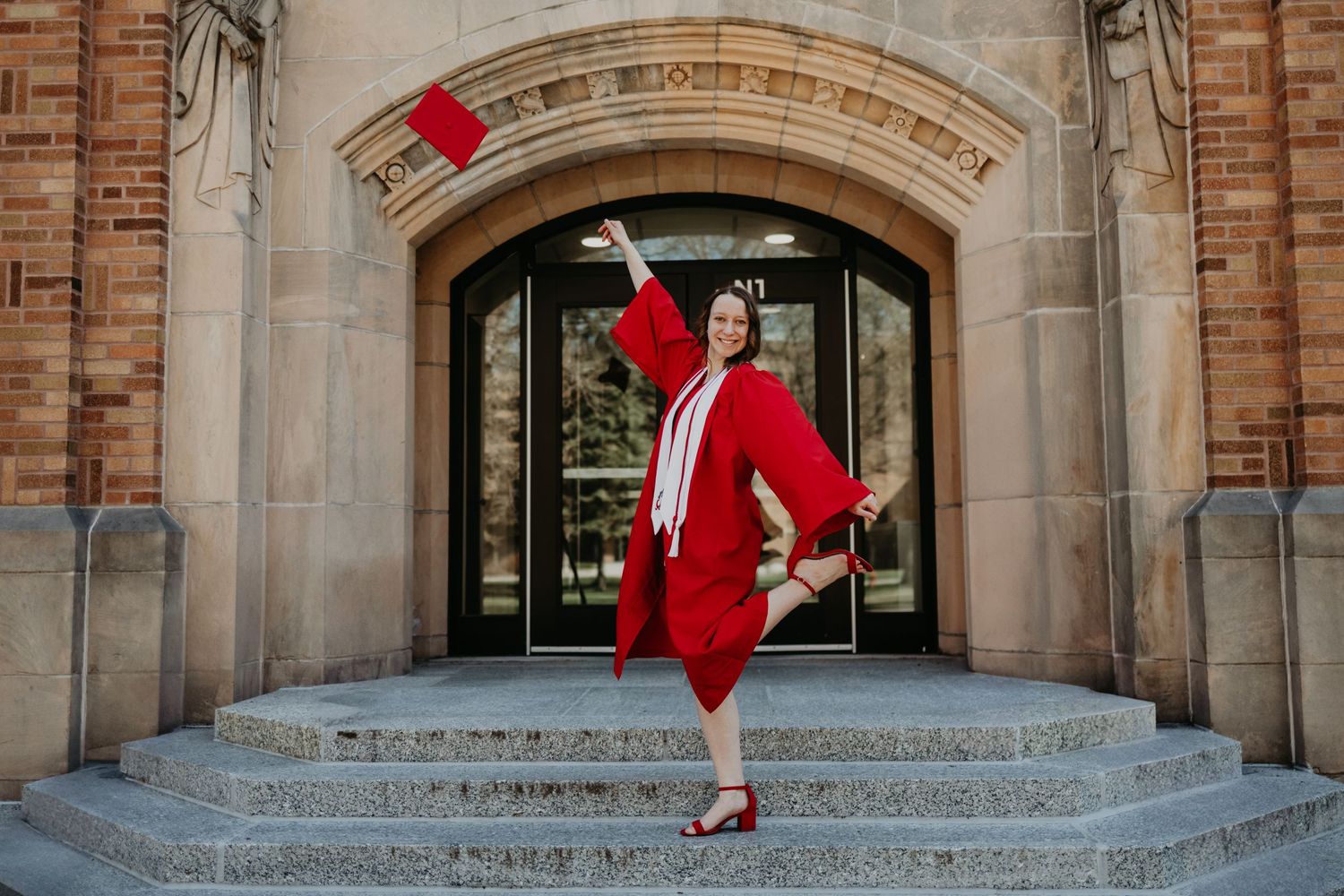 Graduate in red cap and gown posing on stone steps of historic brick building.