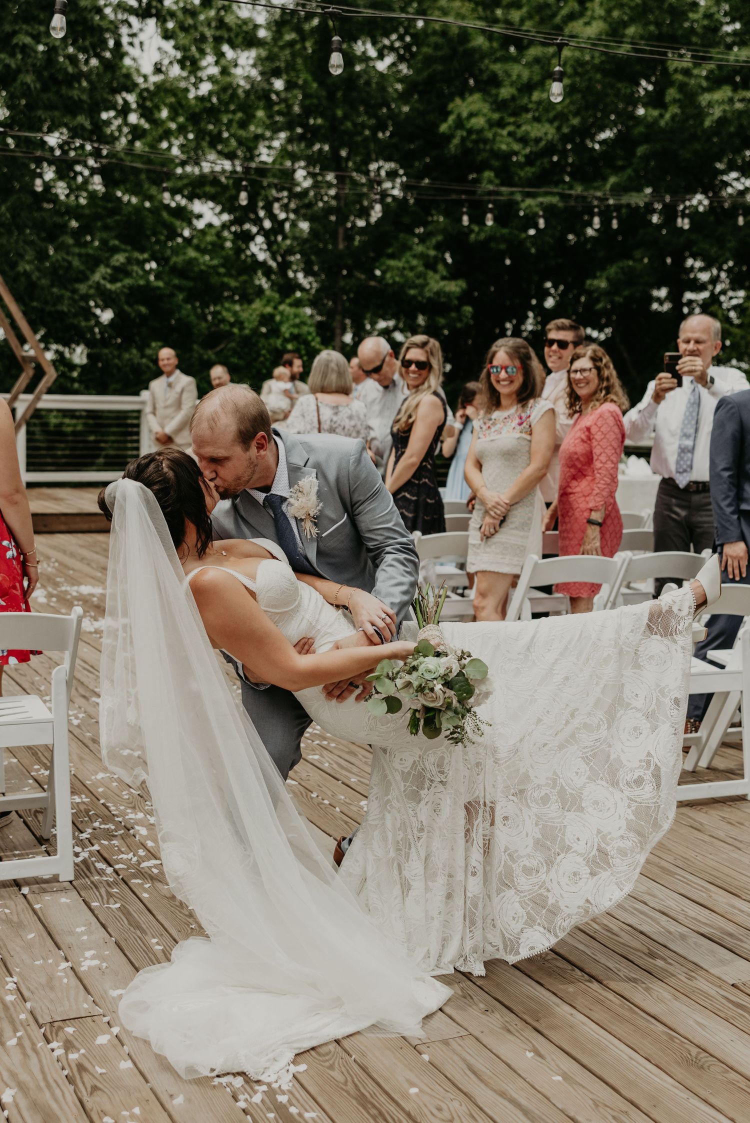 A bride in a flowing lace dress dips dramatically during a dance on a wooden deck.