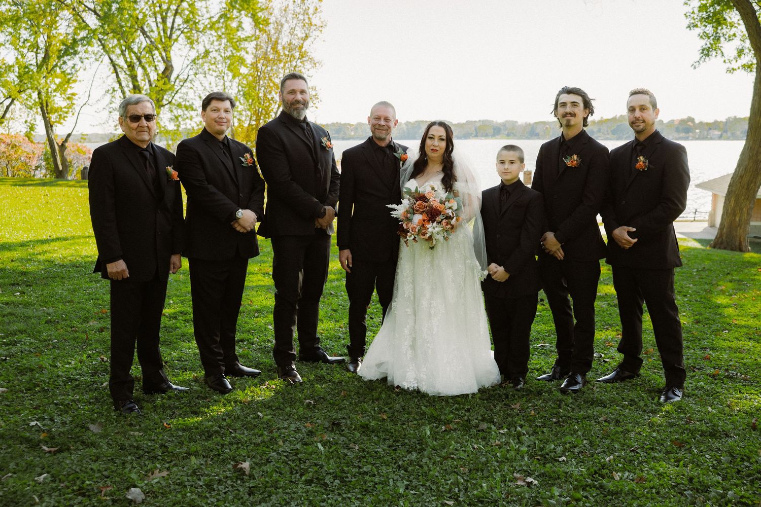 Wedding party in black attire poses on green lawn with river and autumn trees in background.