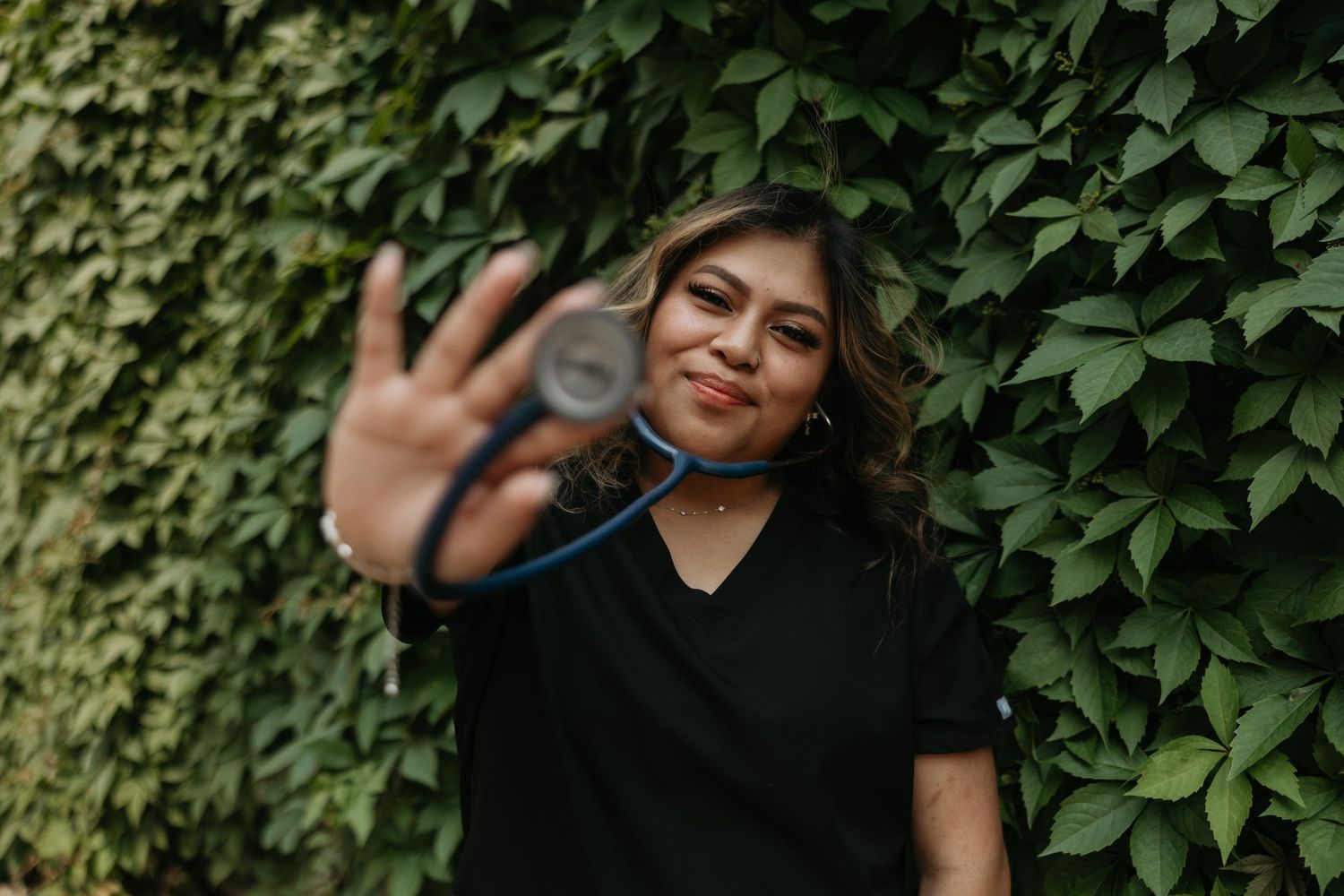 Person in black shirt waving at camera against leafy green wall background.