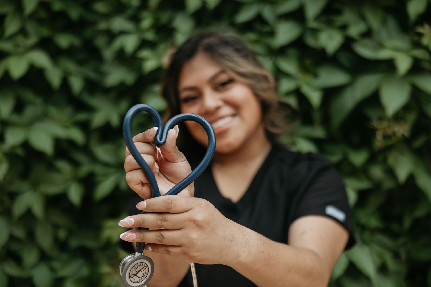 Medical professional forms heart shape with stethoscope against green foliage background.