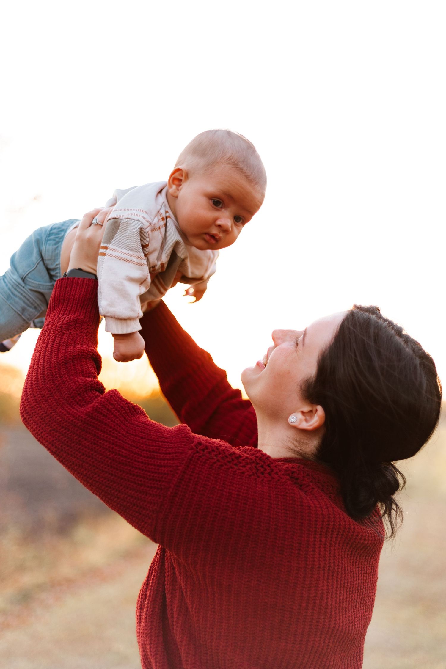 Parent in red sweater lifts and plays with baby against golden sunset backdrop in heartwarming outdoor moment.