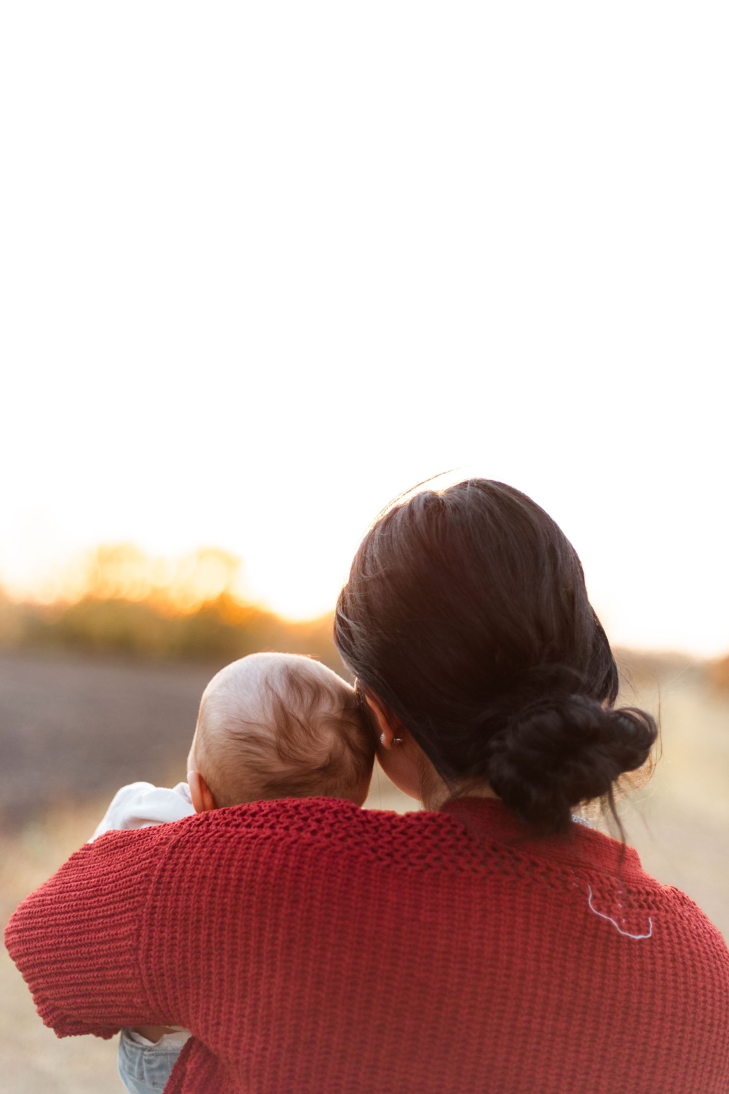 Person in red knit sweater holds baby while watching sunset outdoors in warm evening light.