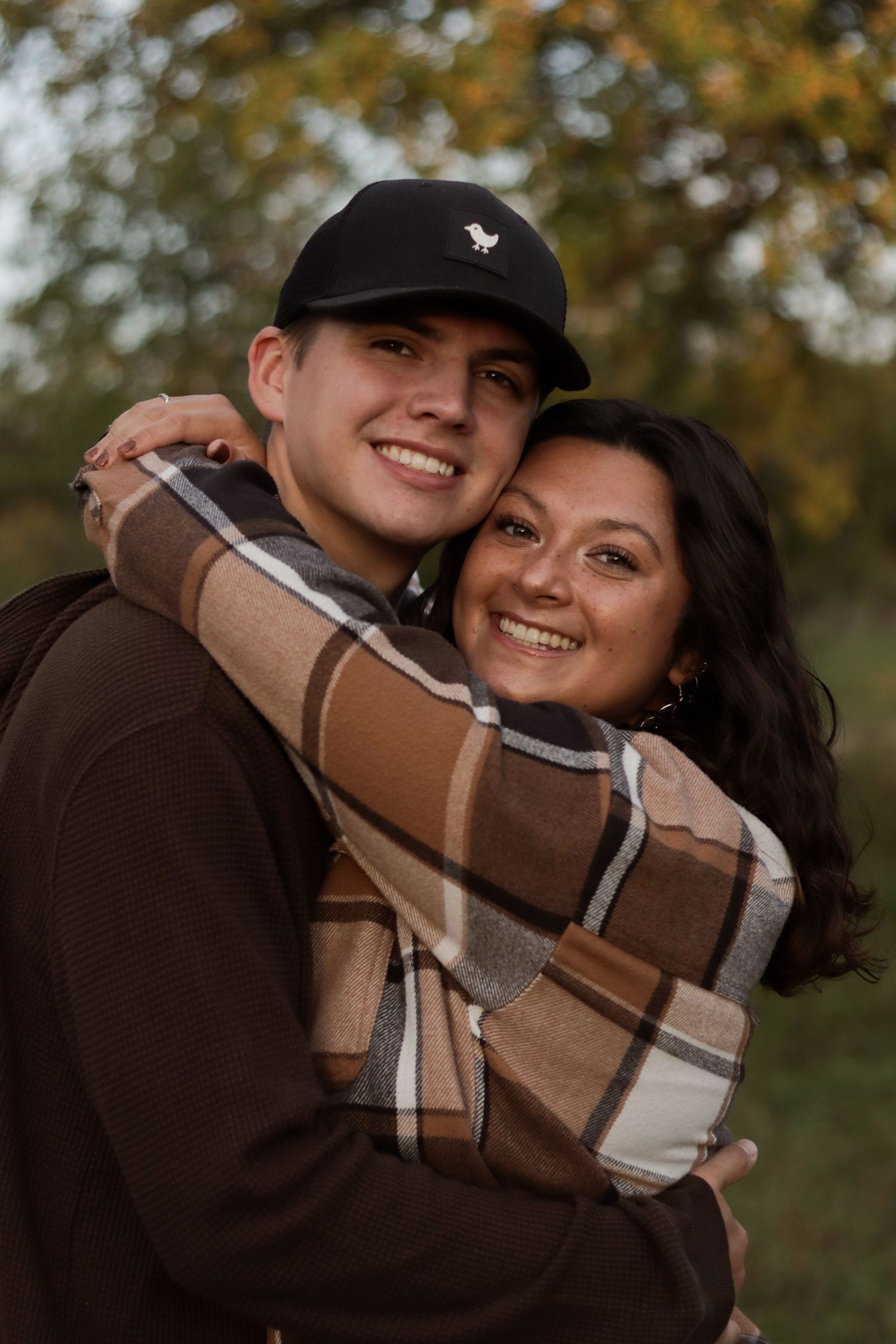 A couple wearing casual fall clothing share a warm embrace and smile outdoors with autumn foliage in the background.