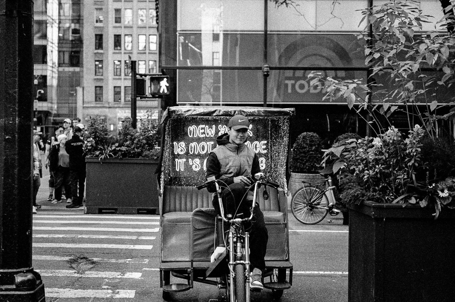 Black and white photo of pedicab driver waiting at intersection in city street.