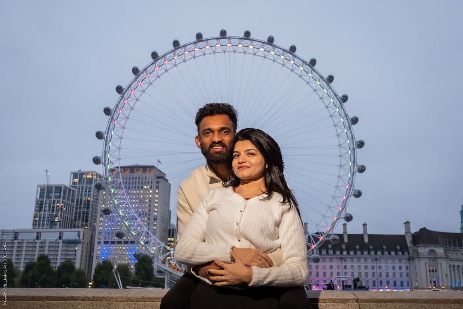 A couple poses romantically in front of the illuminated London Eye at dusk.