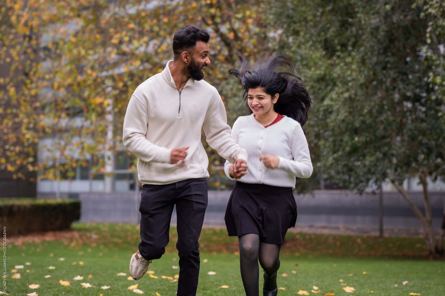 A couple runs playfully through a park on an autumn day.