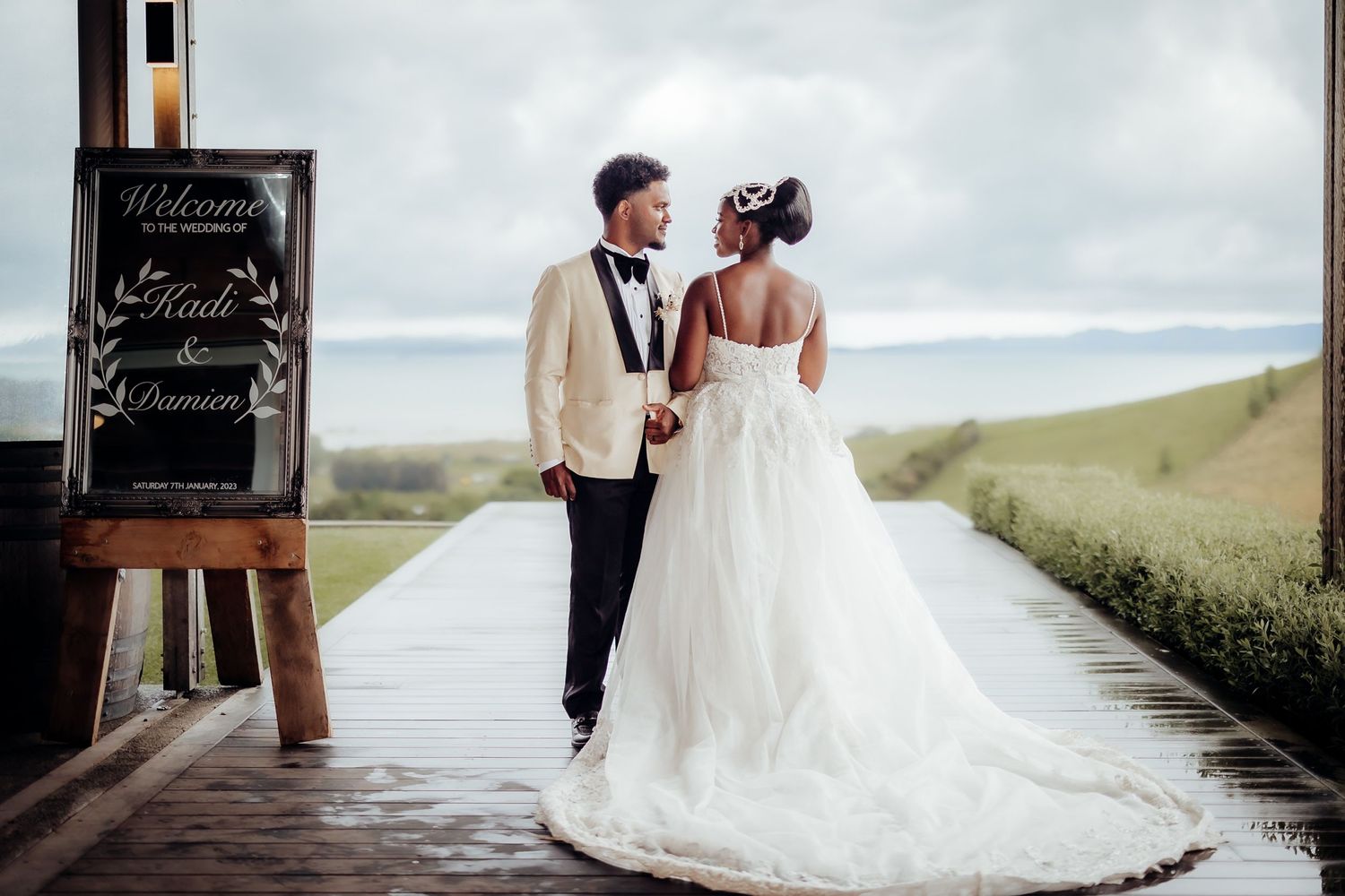 Elegant wedding couple on a wooden deck with a chalkboard sign and scenic backdrop.