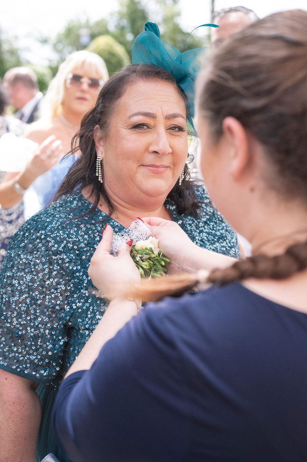 A guest wearing a teal fascinator having a corsage pinned at an outdoor event.