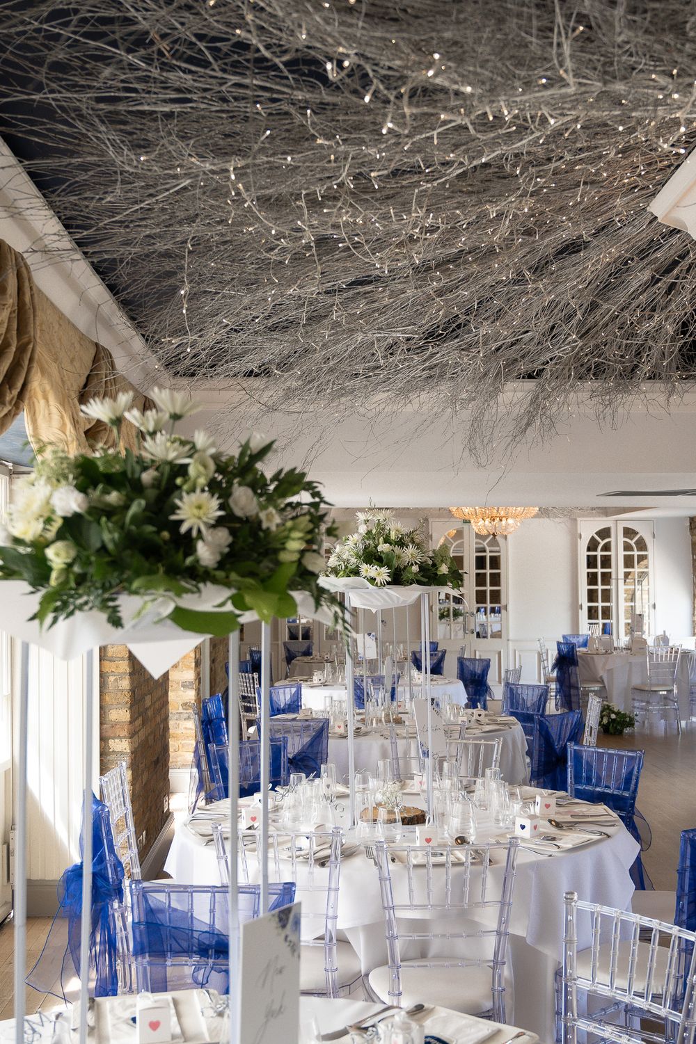 Elegant wedding reception room with blue chair accents and floral centerpieces under a decorated ceiling.